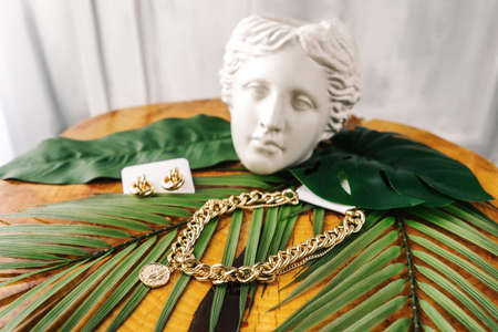 Imitation jewelry, golden bijouterie chain and earrings on leaves on wooden table. White gypsum female head on background like decoration.の写真素材