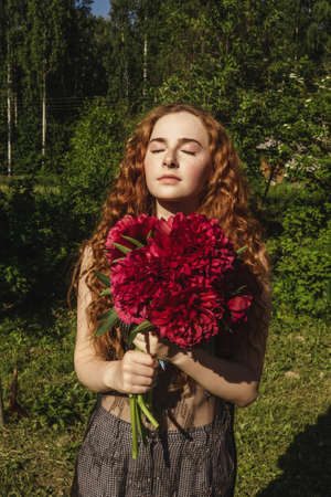 Young beautiful curly ginger woman holds pink peonies in her hands. Green natural background, garden.の写真素材