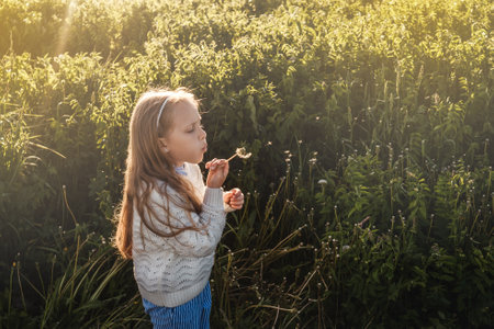 A girl blows on a dandelion in a field. Houses on background.