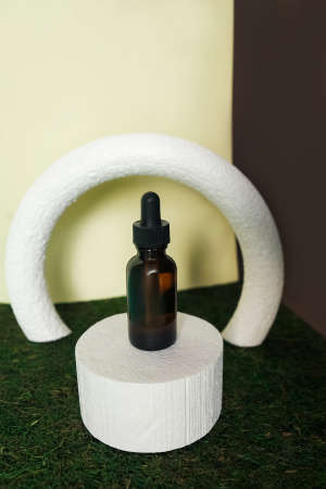 Blank amber glass face serum bottle on yellow and brown background. Abstract background with white podium and arch on green moss floor.の写真素材