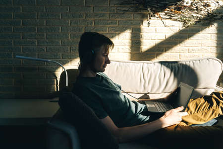 Young man work with laptop on sofa at home. Brick wall and natural pannel on background, sunny day. Man profile in dark.の写真素材