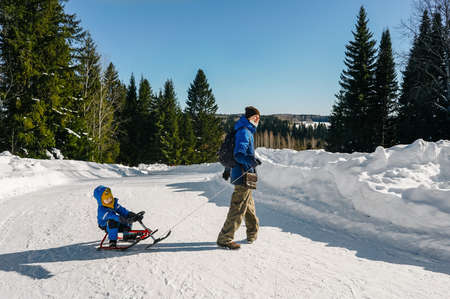 A man run and rolls a small child boy on a snowmobile through the snow in forest road. Front view.の写真素材