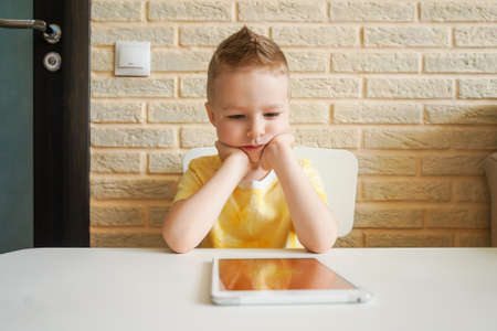 Little sad boy is playing with digital tablet at home. Boy is sitting at the table.の写真素材