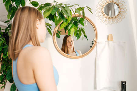 Portrait of beautiful young woman in blue sportswear watch in mirror in morning. Green plant in interior.の写真素材
