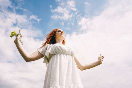 Young beautiful woman with flowers in white dress on sky background. Freedom and piece concept....