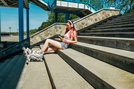 A woman with pink hair and roller skates sits on the steps and listening to music in headphones.の写真素材
