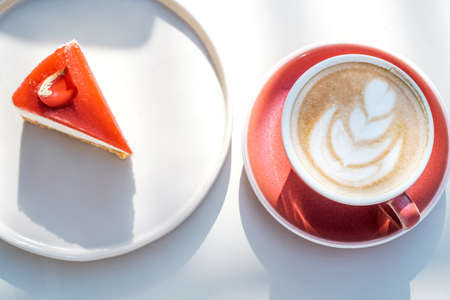 Cup of latte coffee with cheesecake on white table outside. Red cup and red cake. Sunny day. Top view.の写真素材