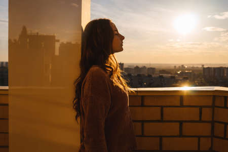 Thoughtful woman with long hair on balcony, city on background.