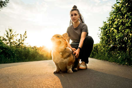 Young happy woman with dreadlocks walk with small dog corgi in park. Sunset light.の写真素材