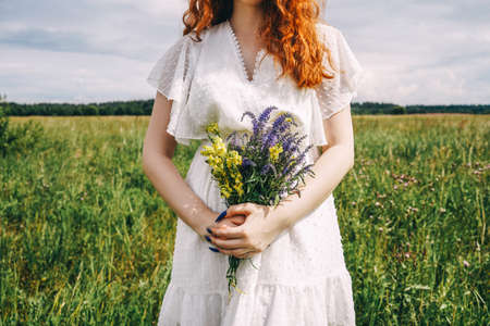 Young beautiful curly ginger woman holds flowers in her hands. Field and sky on background.の写真素材