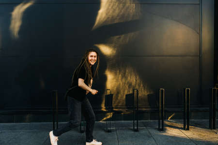 Young happy woman with dreadlocks in park. Modern building on background. Black wall with sun glare.の写真素材