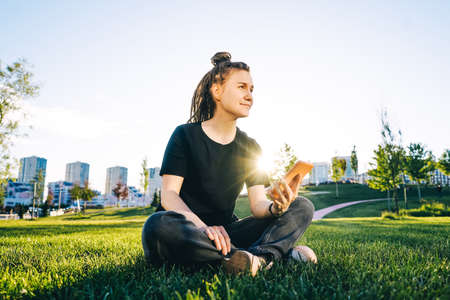 Young happy woman with dreadlocks with mobile phone sitting on green grass in park.の写真素材