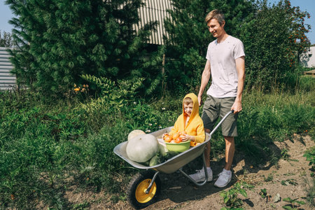 Cute boy with halloween make-up sitting in wheelbarrow with harvest. Father rolls a cart with a baby and vegetables.の写真素材