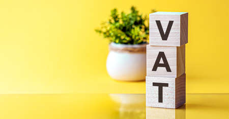 VAT-text on wooden cubes, on a blue background. In the background, a green plant in a white potの写真素材