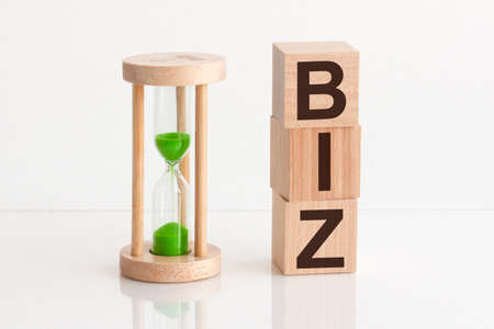 Close-up of an hourglass next to wooden blocks with the text BIZ. biz - text in wooden building blocks, white backgrounds.の写真素材