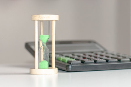 Black calculator and hourglass with green sand close-up stand on a white wooden table, business...