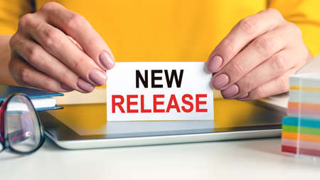 new release written on a white business card in a woman's hands. Yellow background. Glasses, tablet...