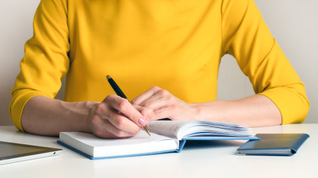 Close-up image of a woman in a yellow dress sitting at an office desk and writing something in a white blank notebook. Selective focus. Can be used for business, marketing, education concept.の写真素材
