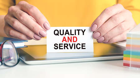 Quality and service is written on a white business card in a woman's hands. Yellow background. Glasses, tablet and block with multi-colored paper for notes. Can be used for business, advertising concept.の写真素材