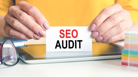 seo audit is written on a white business card in a woman's hands. Yellow background. Glasses, tablet and block with multi-colored paper for notes. Can be used for business, advertising concept.の写真素材