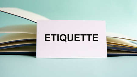 A white business card with ETIQUETTE text stands on a desk against the background of an open diary. Unfocused.の写真素材