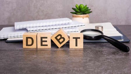 the word Debt is written on wooden cubes on a gray background. close-up of wooden elements, magnifying glass, paper documents and coins on a notebookの写真素材