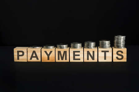 PAYMENTS text written on wooden block with stacked coins on black background, business concept