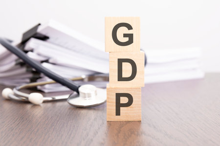 'GDP - Gross Domestic Product' is written on wooden block near a stethoscope on a gray backgroundの写真素材
