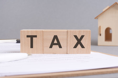 TAX text made of wooden cubes on gray background with a small wooden model houseの写真素材