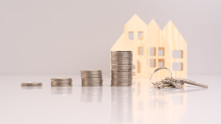 the key is placed next to a stack of coins in front of the small models homes on gray background. wooden model home with key and coins - real estate investment conceptの写真素材