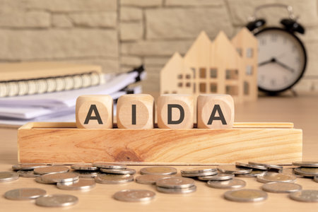 Wooden blocks with AIDA (Attention, Interest, Desire, Action) on a stand, surrounded by coins. In the background, documents, a house model, and a clock create a business and financial atmosphereの写真素材