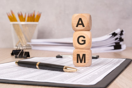 wooden cubes stacked to form 'AGM' on a desk with financial reports, a pen, and office supplies. represents an annual general meeting, corporate governance, financial review, and business strategy.の写真素材
