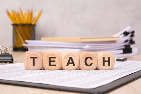 wooden blocks spelling TEACH placed on a clipboard with documents. In the background, pencils and...