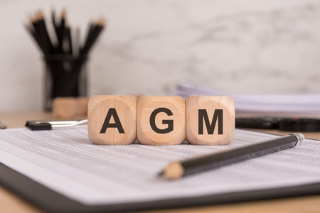 three wooden cubes with the letters AGM on a desk with documents, a pencil, and office supplies,...