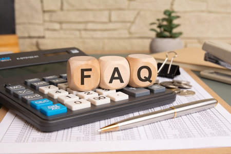 close-up of wooden cubes spelling FAQ on a calculator with financial documents, coins, pen, paper clip, and a potted plant. business office desk concept with financial themeの写真素材