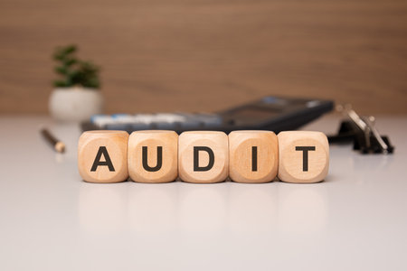 wooden cubes aligned to form the word AUDIT on a white table, with a pen, documents, and calculator in the background.の写真素材
