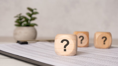 Three wooden cubes with question marks placed on a clipboard with printed spreadsheet. Minimal office scene with soft light and blurred plant in the background. Concept of doubt or inquiry.の写真素材