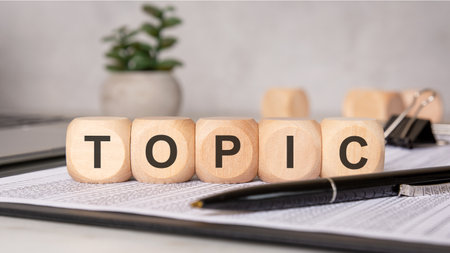 Wooden blocks forming the word TOPIC on financial papers with pen, binder clip and blurred plant, representing content creation, planning, office work or documentationの写真素材