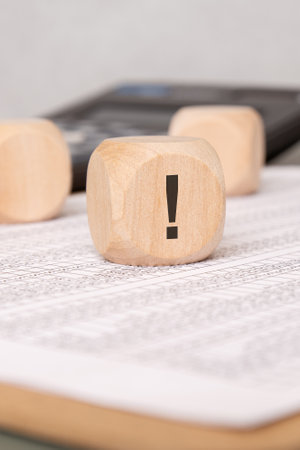 Close up of a wooden block featuring a bold exclamation mark set on a printed finance sheet. Blurred calculator and cubes in background symbolize warning, risk or urgent attention in business analytics.の写真素材