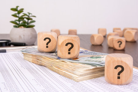 Wooden cubes with question marks placed on a stack of hundred-dollar bills and financial spreadsheets. Concept of financial uncertainty.の写真素材