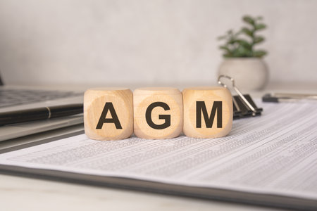 Three wooden cubes with the letters AGM placed on financial documents, next to a laptop, pen, and binder clips - symbolizing an Annual General Meeting in a corporate or business settingの写真素材