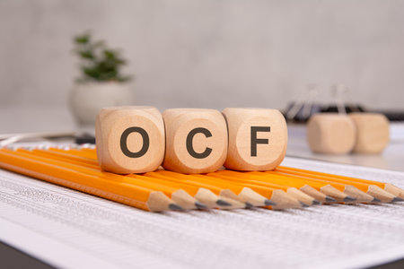 OCF wooden cubes representing Operating Cash Flow, placed on yellow pencils and financial documentsの写真素材