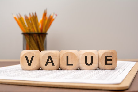 Natural wooden blocks forming the word VALUE placed on a printed financial paper. In the background, out of focus, is a container with pencils. Evokes concepts of business worth, assets, valuation, and economics.の写真素材
