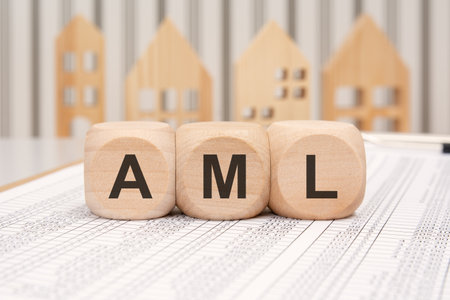 Wooden cubes with AML abbreviation (Anti-Money Laundering) on financial spreadsheet with small wooden houses in background, symbolizing compliance, regulation, and financial crime prevention in banking and real estate sectorsの写真素材