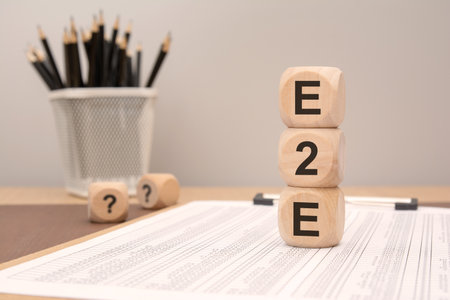 A vertical stack of three wooden cubes printed with E 2 E stands on a document filled with financial data pencils in a holder blur softly in the background. Symbolizes end-to-end workflows, business continuity, and integrated process management.の写真素材