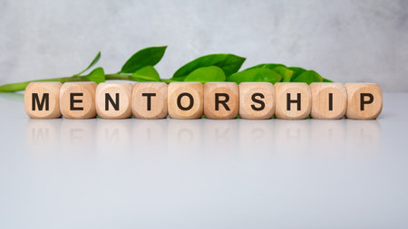 Wooden cubes spelling mentorship on a white glossy surface with green leaves in background. Concept of guidance, leadership, education, support, and personal or professional growth.の写真素材