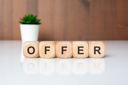 Word OFFER made of wooden cubes on a white glossy table with a blurred plant in background. Minimalistic composition symbolizing business proposal, promotion, or opportunity.の写真素材