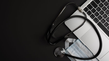 Minimalist top view flat lay of a stethoscope, medical mask, and laptop computer on a dark textured surface. Concept of modern medical technology, online healthcare services, digital diagnosis, and remote patient monitoring. Plenty of negative copy space on the left.の写真素材