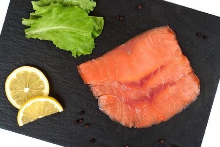 Sliced salmon fillet on the black shale board, with lemon and salad. On white background