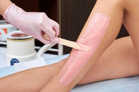 The beautician is preparing for depilation and applying the cream with wax stick on the female legs. Cosmetology procedures at homeの写真素材